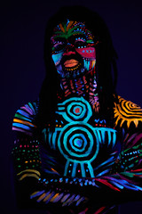 portrait of fashionable male model with fluorescent UV body art posing in dark space, male with dreadlocks, various ethnic colorful prints on naked skin