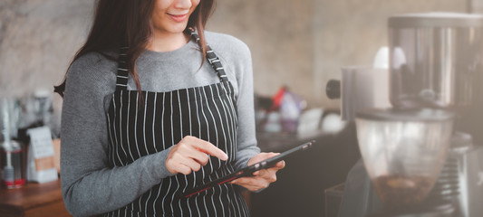 young baristas using tablet smiling and looking at her touchscreen smartphone get orders from online shopping while standing at coffee bar.for banner, web page or cover template