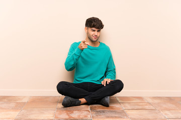 Young handsome man sitting on the floor points finger at you