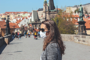 Young woman with sunglasses and medical face mask photographed on the Charles Bridge in Prague, Czech Republic. Blurred old town in the background. Travelling, tourism during coronavirus. COVID-19