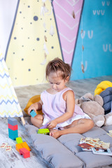 Fototapeta premium Little girl plays wigwam with colorful toy bricks and builds tower. Happy baby plays in tent in children's room with educational toys. baby plays in kindergarten. childhood concept, child development 