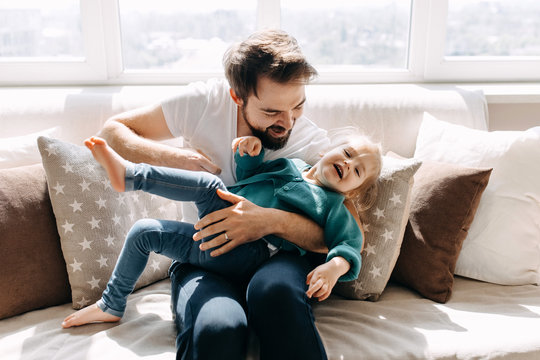 Father and little daughter playing together, sitting on the sofa by the window.