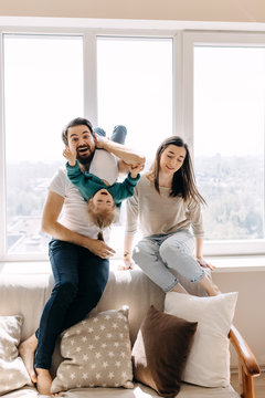 Family of three, father, mother and little daughter playing together, sitting on the sofa by the window.
