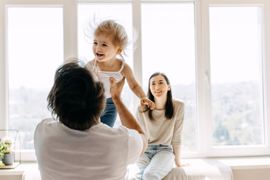 Family of three, father, mother and little daughter playing together, sitting on the sofa by the window.