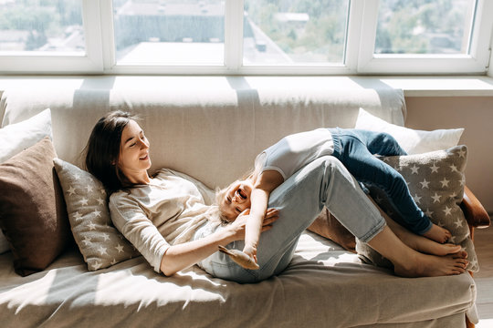 Mother and little daughter playing together, lying on the sofa by the window.