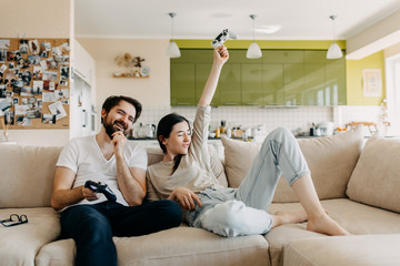 Young couple of man and woman playing video game on a console, sitting on couch in living room, girl winning.