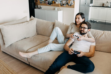 Young couple of man and woman playing video game on a console, sitting on couch in living room, hugging.