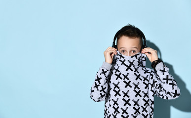 Teenage kid in headphones and black bracelet. He is hiding his mouth in stylish hoodie while posing against blue background