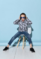 Teenager in headphones, sunglasses, casual clothes. Looking wondered, listening to music. Sitting on white chair. Blue background