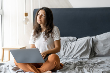 Beautiful young brunette girl with a laptop sitting on the bed. Stylish modern interior. A cozy workplace. Shopping on the Internet