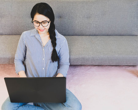 selective focus young beautiful asian woman wore blue striped shirt and jean,sitting on pink carpet by the sofa,work online lifestyle relax at home working with laptop,work from home concept