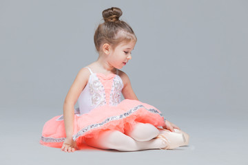Cute charming ballerina little girl in pink tutu is sitting on the floor trying to put on ballet...