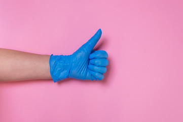 Doctor hand in blue disposable medical glove showing thumbs up on pink background. Gesture of...