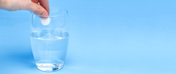 Effervescent instant tablet in a hand over a glass of water on a blue background. Banner with copy space. Treatment, vitamin intake during the pandemic of the coronovirus