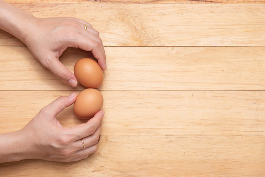 Top View Of Two Hand Picking Up Two Raw Chicken Eggs On A Light Brown Wooden Background, Copy Space