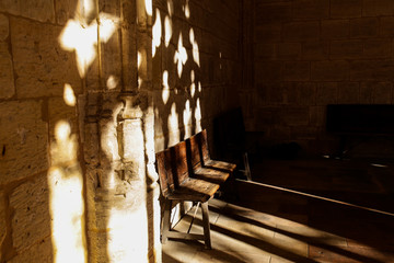 Fototapeta na wymiar A wooden bench stands in the corner of the annex to the old stone church and the bright day sun with its rays through the openwork lattice draws elaborate patterns on the bench and the wall above it