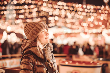 A young woman tourist stands smiling on the street of a night city during the Christmas and New Year holidays. In the background are the very blurry lights of festive illuminations. Tinted.