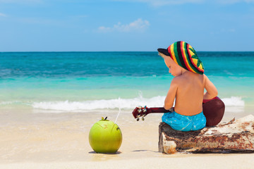 Little baby in rasta hat play reggae music on Hawaiian ukulele, enjoy relaxing on ocean beach. Kids...