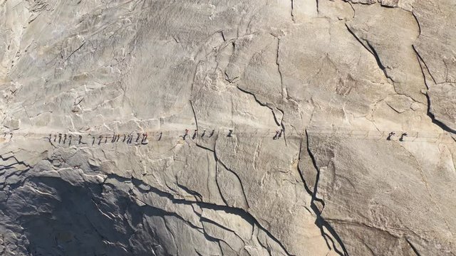 Top aerial footage of a group of tourists climbing a specially equipped trail which goes up a smooth stone slope to the top of the famous Half Dome mountain, located in Yosemite National Park, USA. 4K