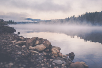 Dramatic view of a calm mountain lake covered by fog just before sunrise in Smida village. The quiet nature with fog above the lake surrounded by fir and spruce forest in the pleasant morning light.