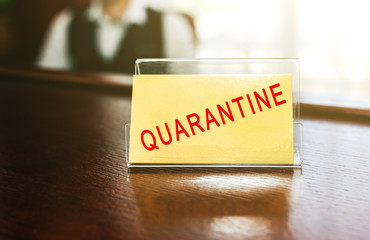 Quarantine text on business card at the holder on the wooden reception at the hotel. The administrator is sitting on background at the desk. Coronavirus people isolation - obrazy, fototapety, plakaty