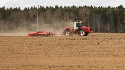 Obraz premium Red big powerful wheeled tractor with large plough ploughs the land in a field at spring day on forest trees backgroung, rural farming landscape