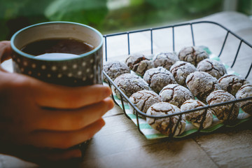 Chocolate crinkle cookies in a basket with a cup of tea hold by hands on a wooden table. Close up view. Landscape format.