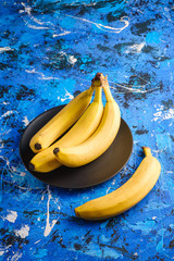 Banana fruits in black plate on blue textured background, angle view copy space