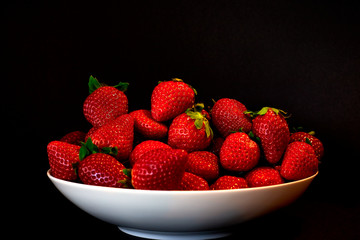 Imagen horizontal de apetitosas fresas rojas, maduras y jugosas con hojas verdes llenando un plato blanco ante un fondo negro.