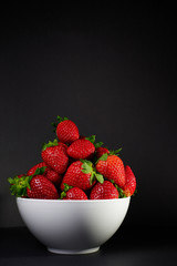 Imagen vertical de apetitosas fresas rojas, maduras, jugosas y apetitosas con hojas verdes llenando un bol blanco ante un fondo negro.