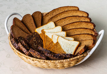 Close-up of pieces of black bread with seeds, grey and two-colored  cutted and served on the wicker dish.  Cream tablecloth with leaves texture.
