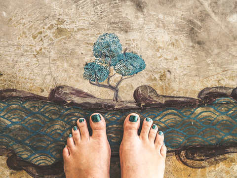 top view of feet with blue painted nails standing on a drawing of a tree and a river on a vintage concrete floor