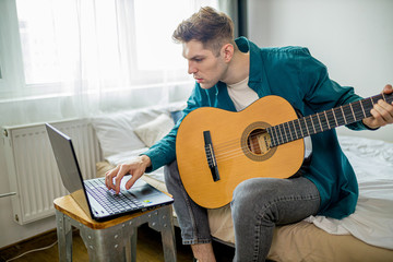 side view on young self-taught caucasian man want to learn new melody for playing guitar at home, man use acoustic guitar, search new online lessons in laptop