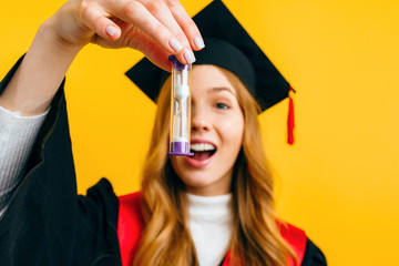 beautiful graduate holds an hourglass in her hand on an isolated yellow background