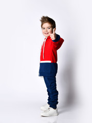 Little blond son in blue and red tracksuit, sneakers. Smiling, showing thumb up, posing sideways...