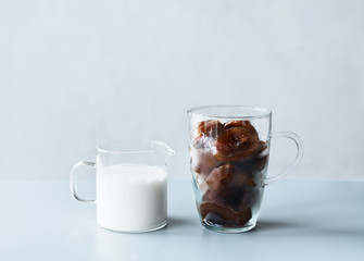 Coffee ice cubes in glass mug and little jug with cream or vegan milk on grey table at wall background. Refreshing summer drinks making ingredients