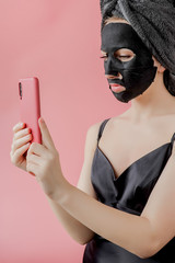 Young woman apply black cosmetic fabric facial mask and phone in hands on pink background. Face peeling mask with charcoal, spa beauty treatment, skincare, cosmetology. Close up