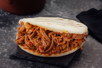 Arepa stuffed with shredded meat on a dark background. (Arepa con carne mechada)