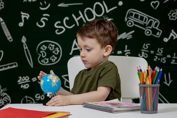 Cute clever boy is sitting at a desk with globe in hand on background with blackboard . Ready for school. Back to school