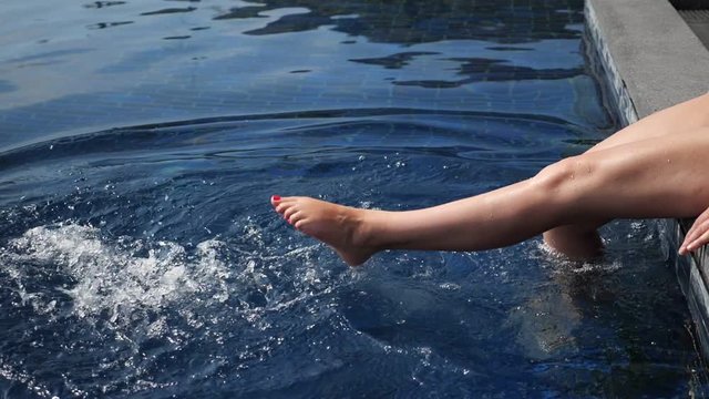 young female legs splashing in blue swimming pool water on summer day slow motion