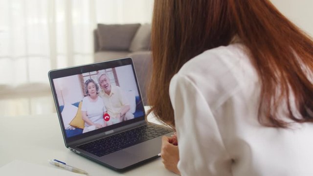 Young Asian business female using laptop video call talking with family dad and mom while working from home at living room. Self-isolation, social distancing, quarantine for coronavirus prevention.