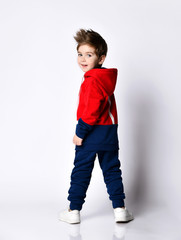 Little blond boy in blue and red tracksuit, sneakers, sunglasses. Smiling turning back, showing thumb up, posing isolated on white