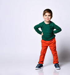 Cute brunet child in green jumper, orange pants, blue sneakers. He put hands on hips and looking at you, posing isolated on white