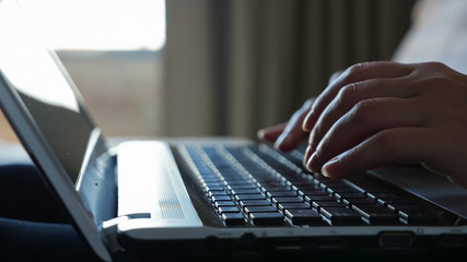 girl freelancer hands type on black laptop keyboard against blurry window and brown curtains in bedroom extreme closeup