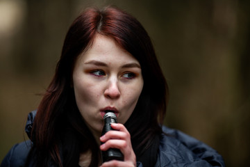 Vape teenager. Young pretty caucasian brunette girl smoking an electronic cigarette on the street in the spring. Deadly bad habit.