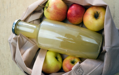 A bottle of apple juice lies in an eco bag with apples on a wooden table.