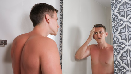handsome guy with bare torso looks at sunburned face and body in mirror standing in hotel bathroom close view