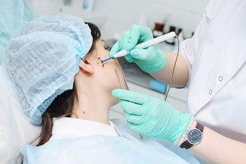 Fototapeta premium A young woman doctor dermatologist surgeon holds a radio wave knife in the background of a young female patient. Removal of moles, warts, rosacea, vascular asterisks, pigmentation.