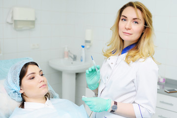 A young woman doctor dermatologist surgeon holds a radio wave knife in the background of a young female patient. Removal of moles, warts, rosacea, vascular asterisks, pigmentation.