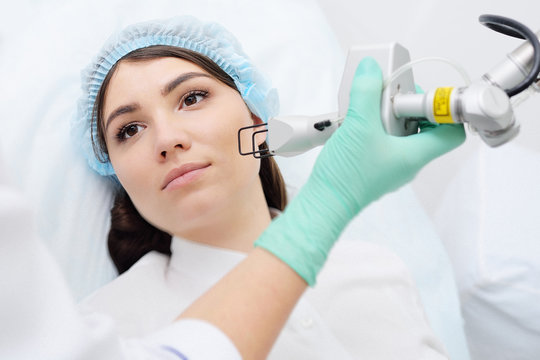 doctor cosmetologist dermatologist conducts a laser skin rejuvenation procedure for a patient-a young pretty woman with a modern fractional laser. Laser cosmetology, removal of scars